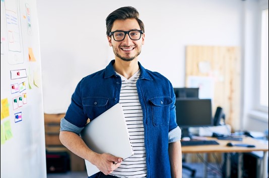 Man holding laptop, smiling, wearing glasses and posing for photo