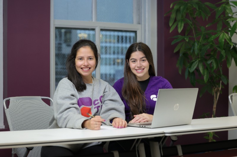 Two women posing with a smile, laptop and pen for a photo